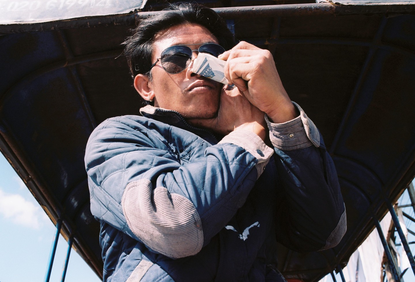 A man wearing sunglasses holds a folded banknote to his nose while sitting in a vehicle.