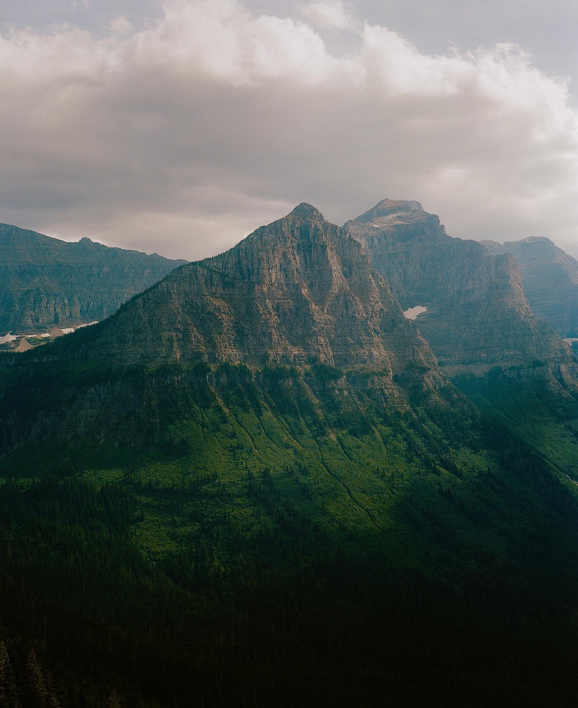 A mountain range with a prominent peak covered in green foliage and dark forest at its base is visible under a cloudy sky.