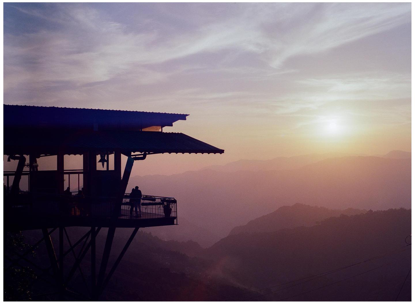 A building with a large deck overlooks a mountain range at sunset.