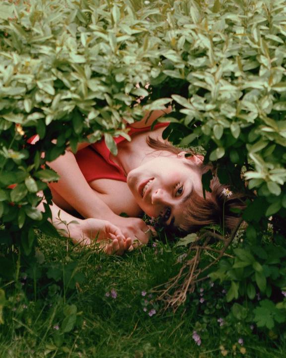 A woman in a red top smiles at the camera while lying in a garden, partially obscured by green foliage.