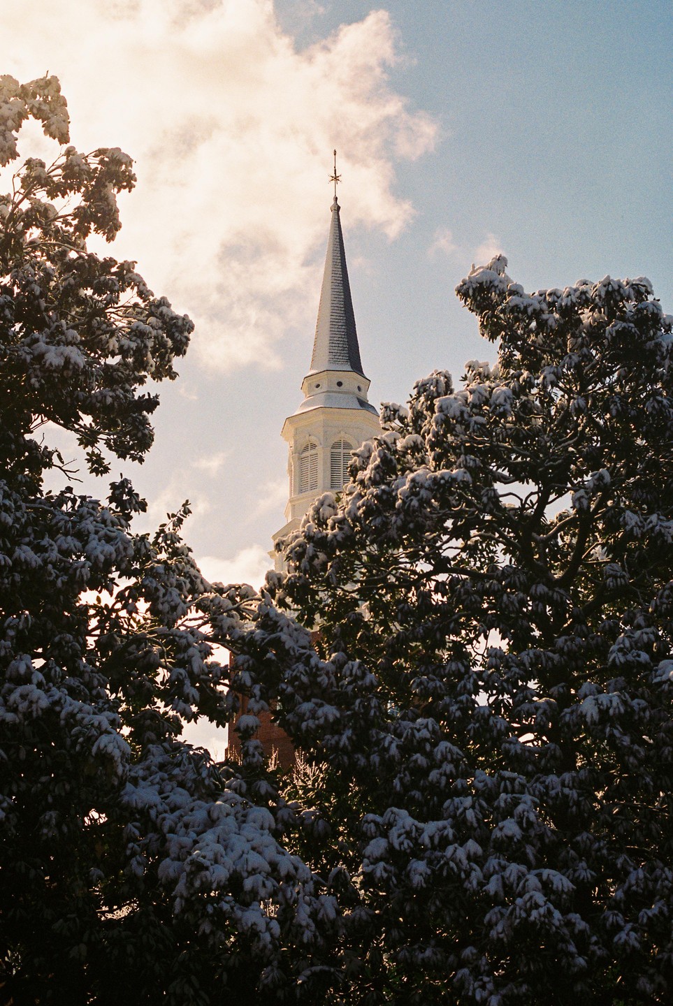 A church steeple rises above snow-covered trees against a cloudy sky.