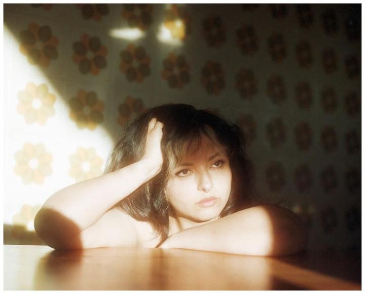 A woman with dark hair and light bangs rests her head on her arm on a wooden table, with a retro floral wallpaper in the background.