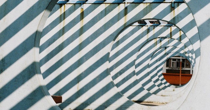 Sunlight casts diagonal shadows through a series of concrete arches, revealing a building in the distance.