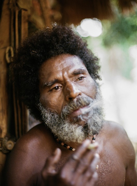 A man with a gray beard and curly hair smokes a cigarette.