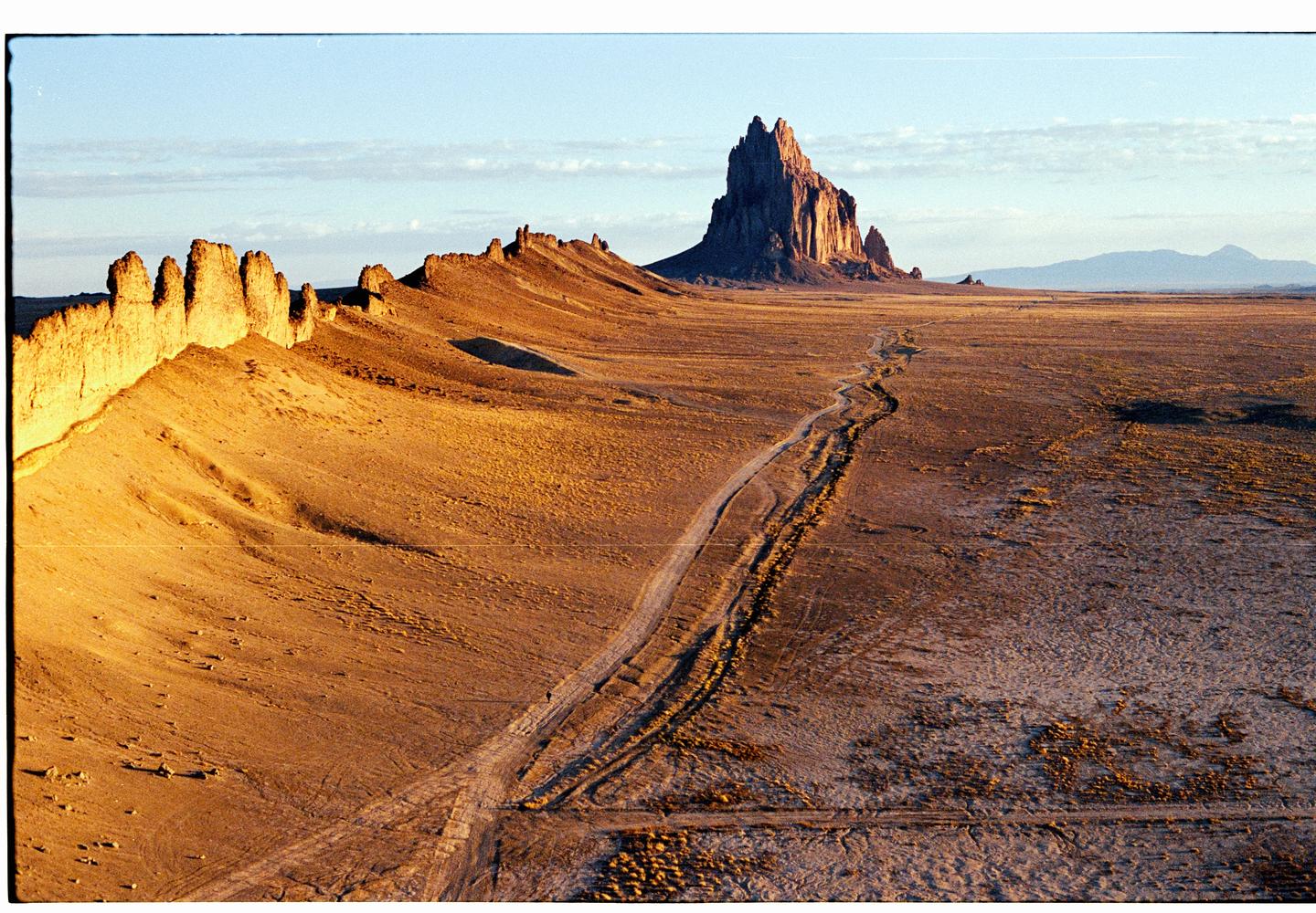 A dirt road leads through a vast desert landscape towards a large rock formation under a clear sky.