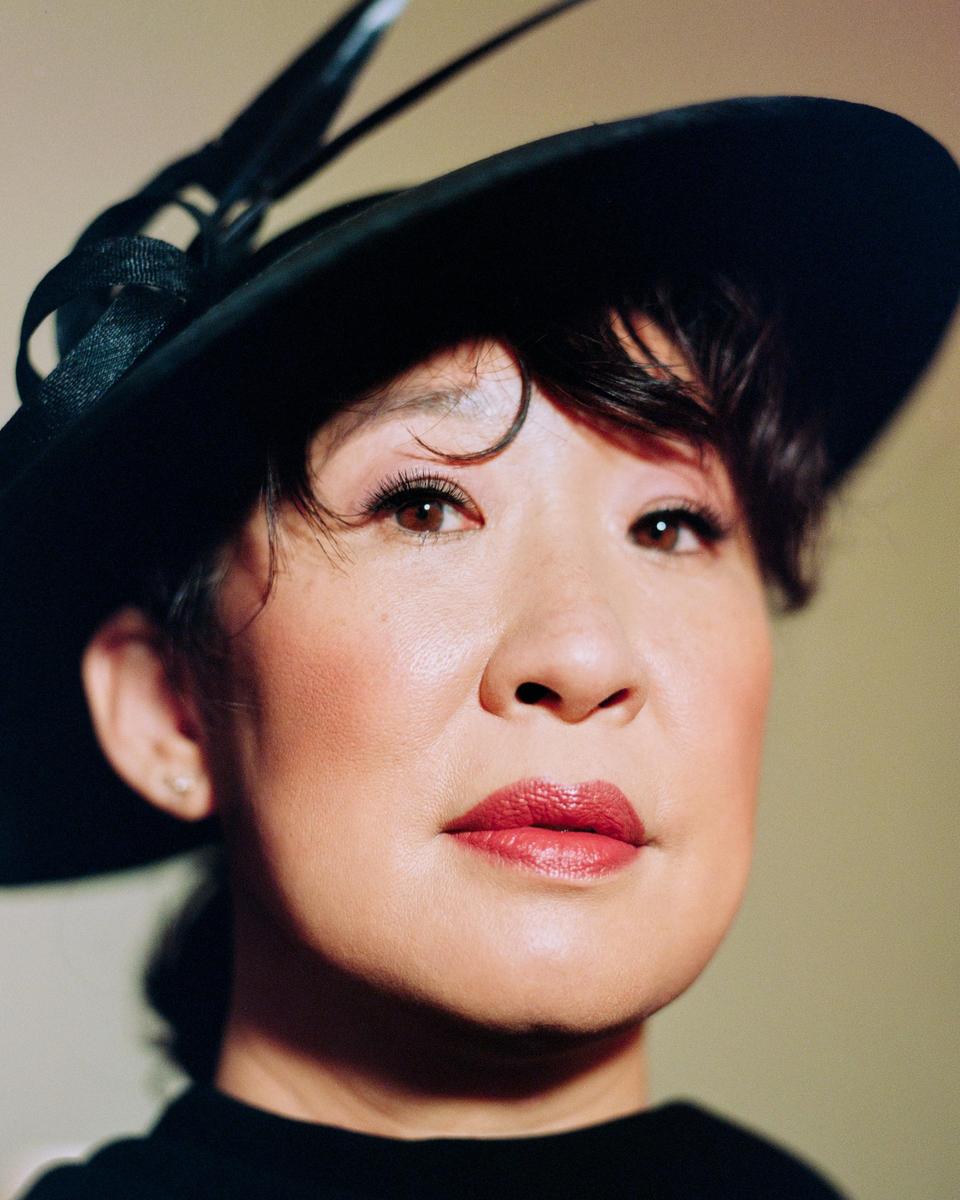 A woman wearing a black hat with feathers looks directly at the viewer.