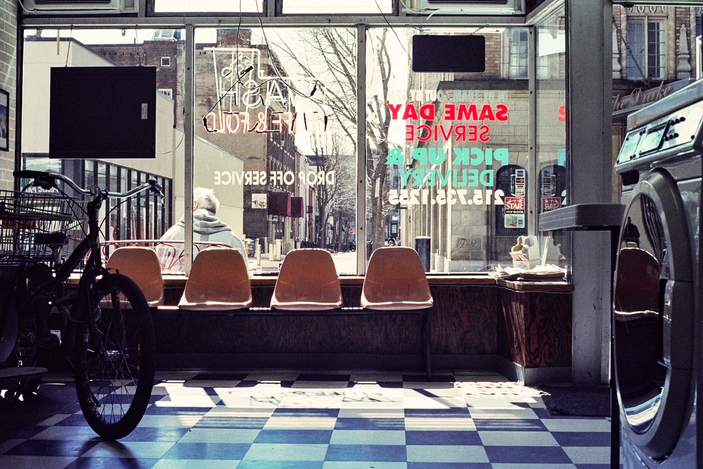 A man sits on a bench inside a laundromat with a bicycle in the foreground and a street visible through the window.