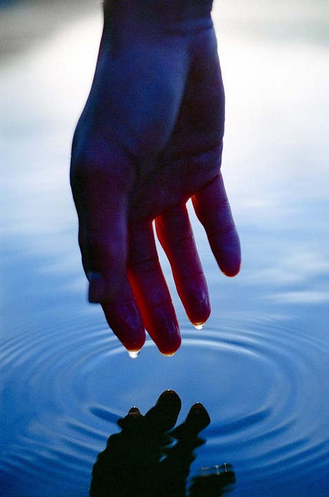 A hand reaches down to touch the surface of blue water, creating ripples.
