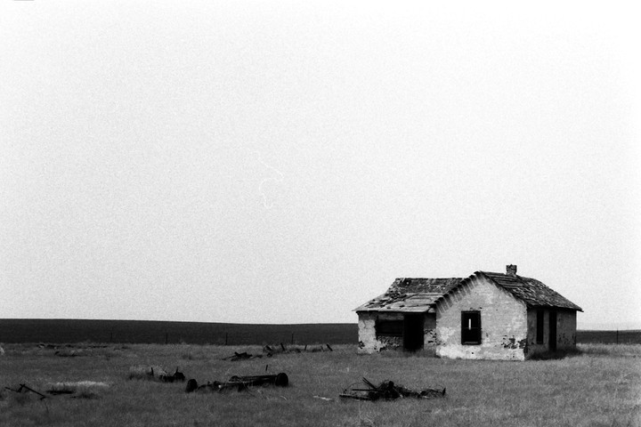 An abandoned house with a damaged roof stands in a grassy field under a bright sky.