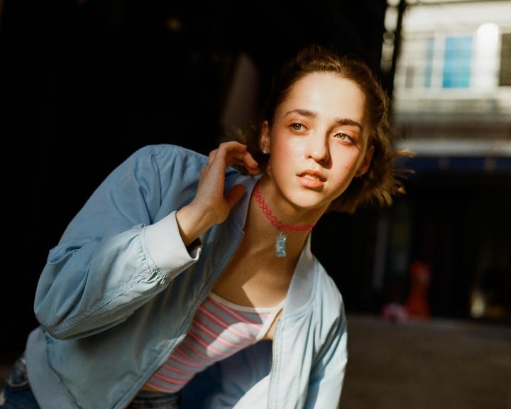 A young woman with a pink choker and a light blue jacket looks away from the camera.