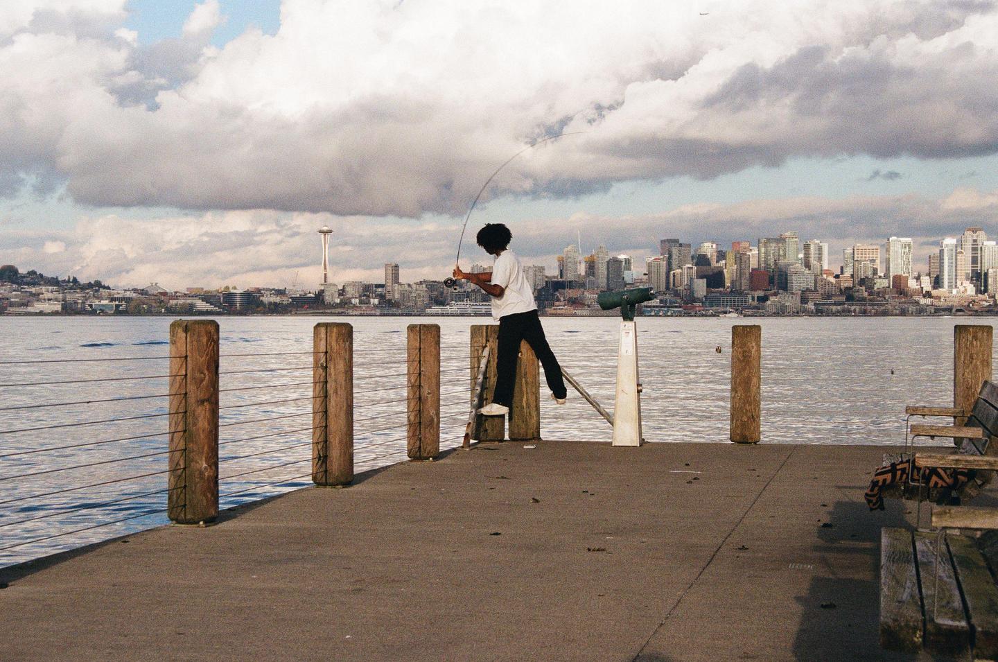 A man stands on a pier holding a fishing rod with the Seattle skyline in the background.