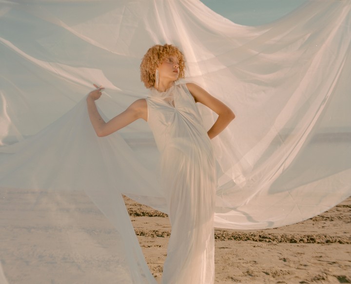 A woman with curly hair poses on a sandy beach, holding a large piece of flowing white fabric.