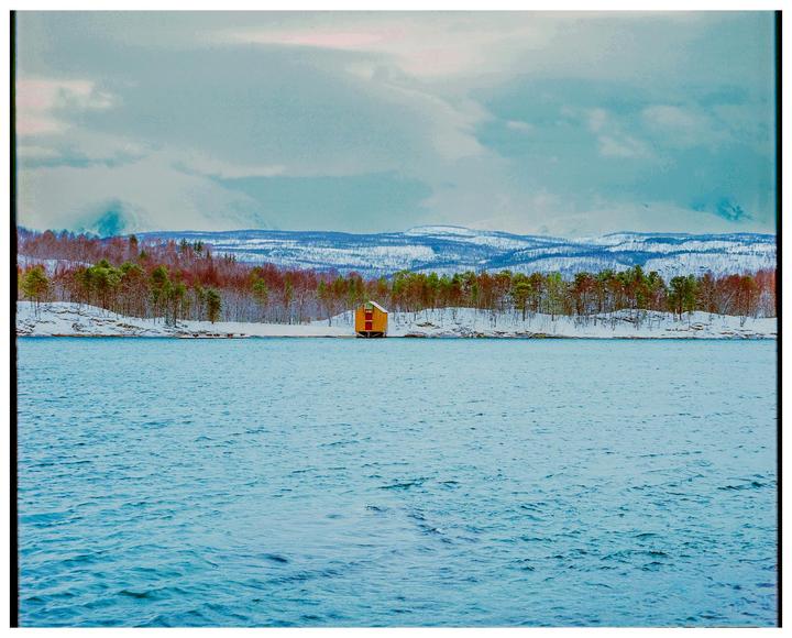 A small yellow house sits on a snowy shoreline with a forest and mountains in the background under a cloudy sky.