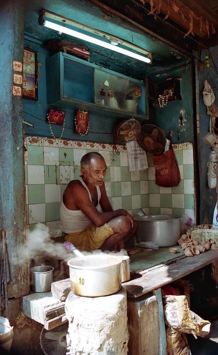 A man squats next to a steaming pot in a small, tiled room with blue walls.