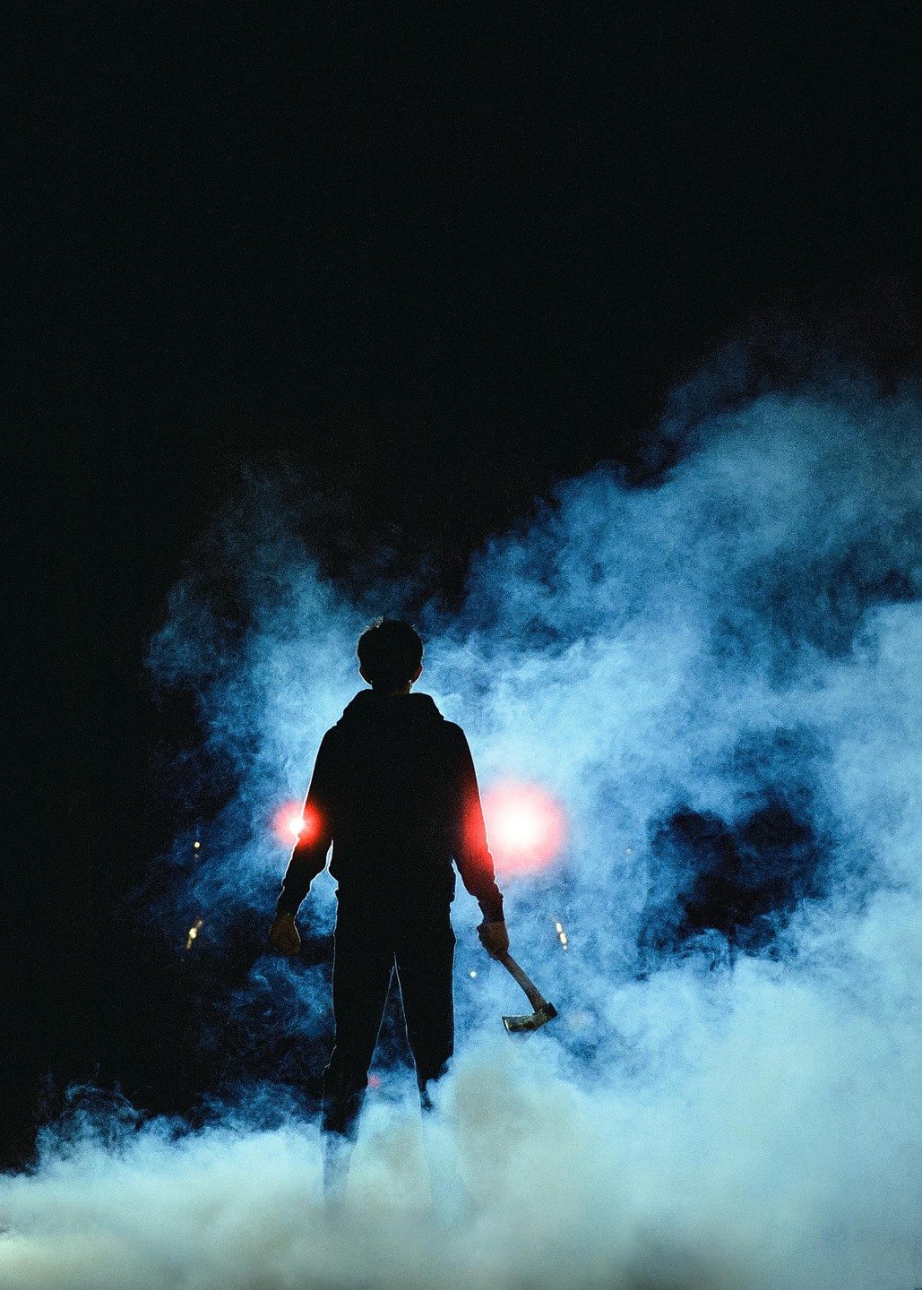 A man holding an axe stands in a cloud of smoke with red lights behind him at night.