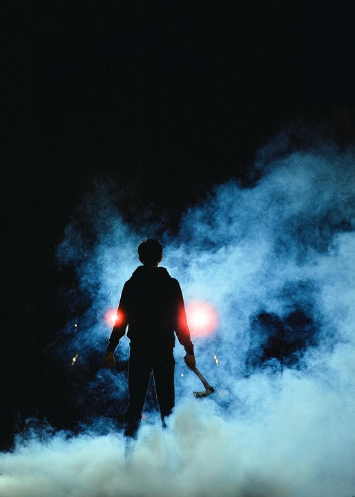 A man holding an axe stands in a cloud of smoke with red lights behind him at night.