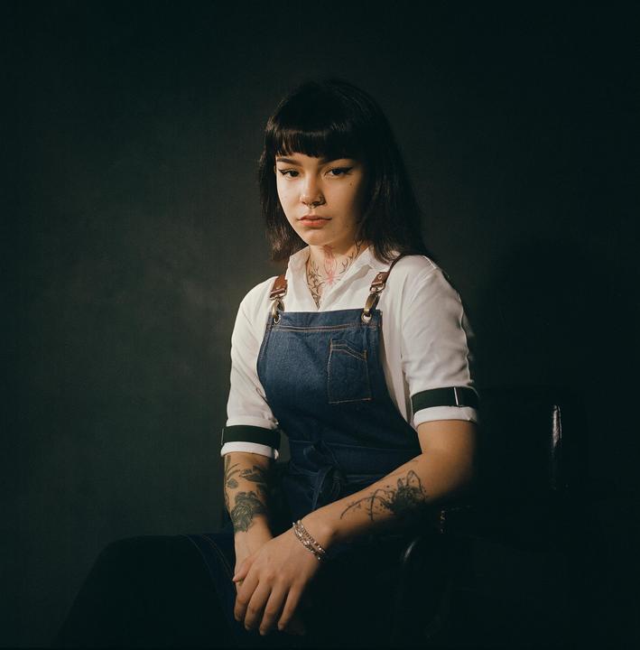 A woman with dark hair and tattoos sits wearing a denim apron over a white shirt.