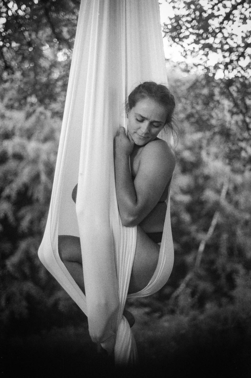 A woman is nestled in a white fabric hammock outdoors, with trees in the background.