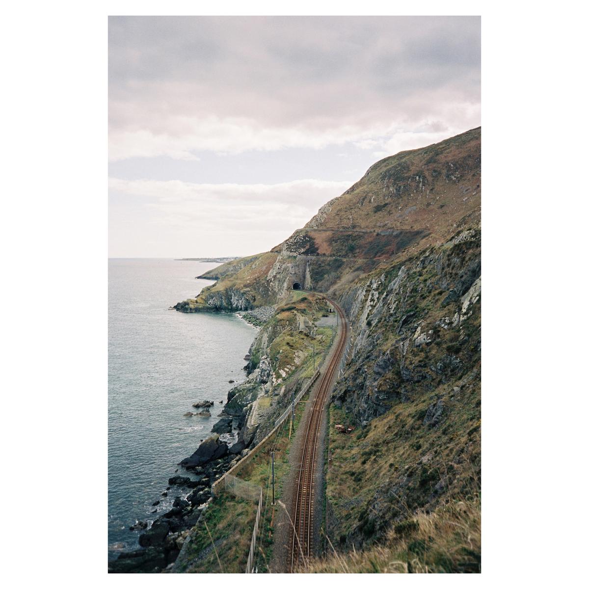 A train track curves along a rocky coastline with a tunnel entrance visible in the cliffside.