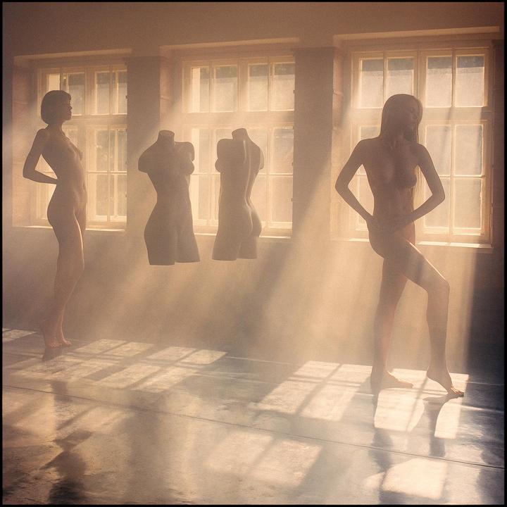 Two nude women and two headless mannequins stand in a hazy room with sunlight streaming through windows.