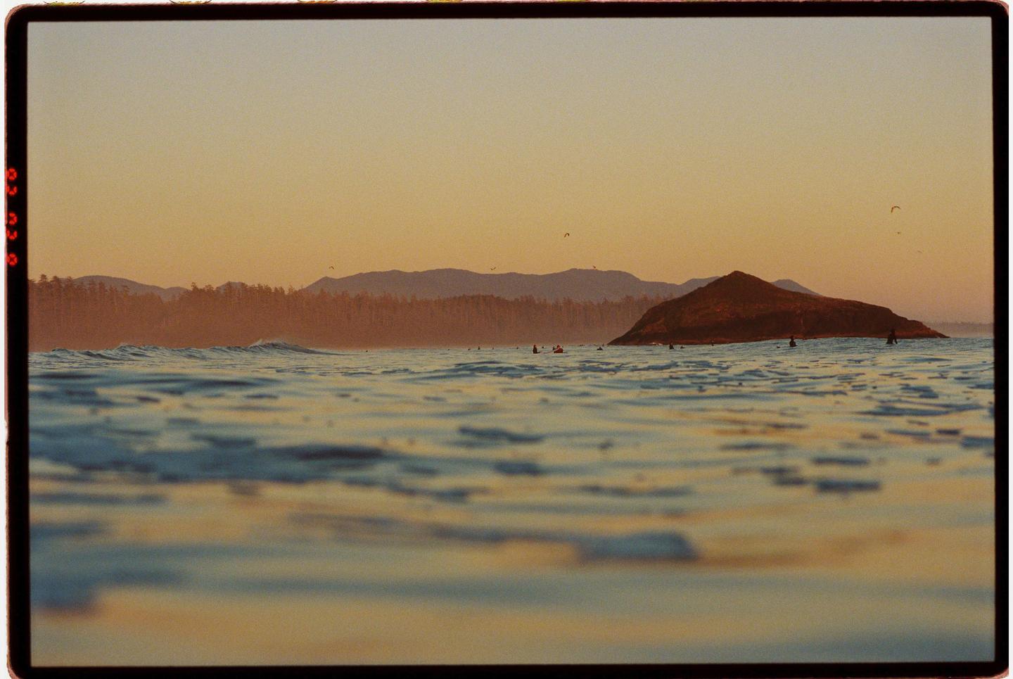 A body of water with small waves is in the foreground, with a small island and mountains in the background under a golden sky.