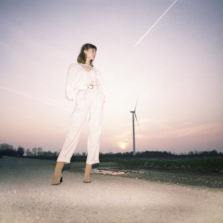 A woman in light-colored clothing stands on a dirt road with a wind turbine and sunset in the background.