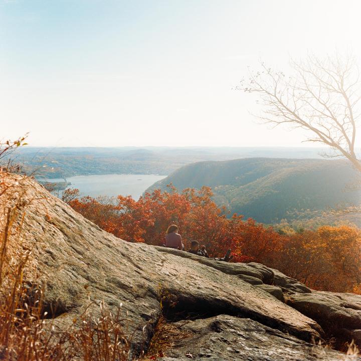 A woman and child sit on a rocky outcrop overlooking a river and mountains covered in autumn foliage.