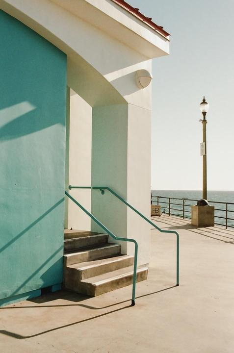 A turquoise wall and white building with stairs and a railing lead to a pier with a lamppost and the ocean in the background.