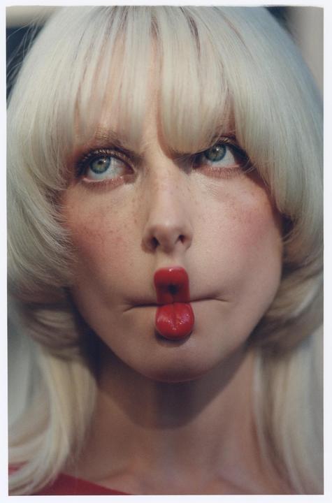 A woman with blonde hair and blue eyes has a red candy between her lips.
