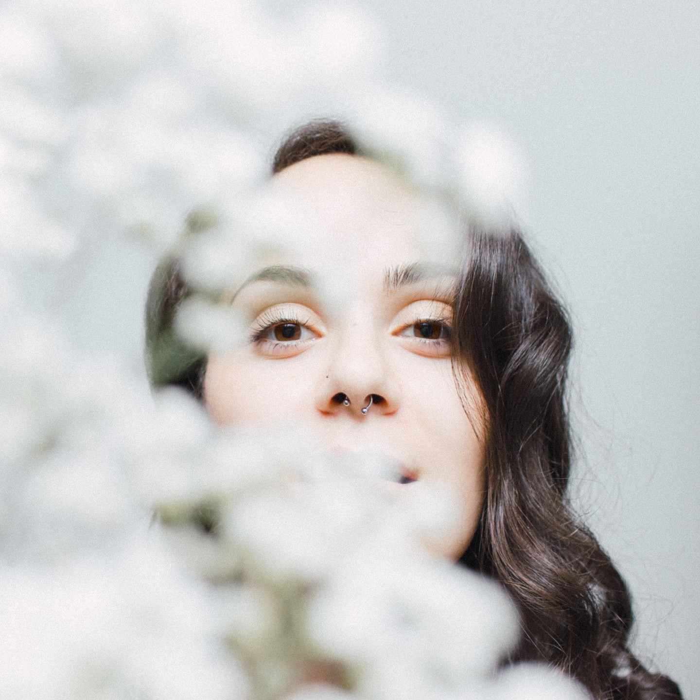 A woman with dark, wavy hair and a septum piercing looks directly at the viewer, partially obscured by white blossoms in the foreground.