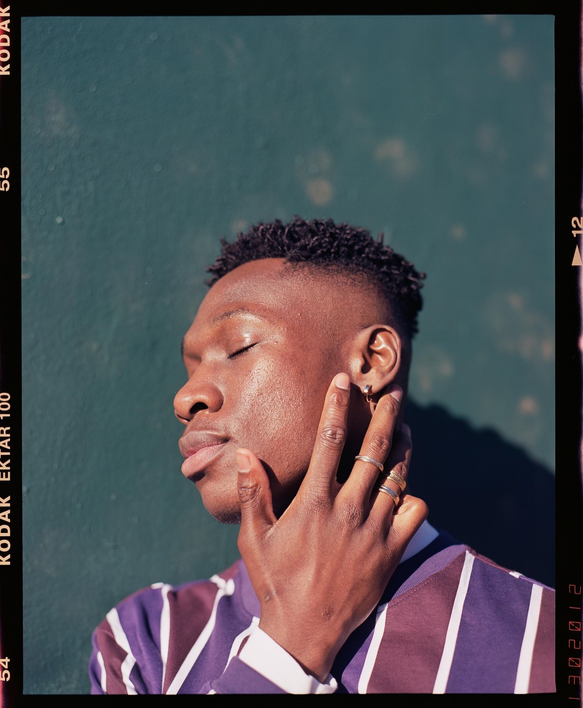 A man with closed eyes and rings on his fingers touches his face, with a green wall in the background.