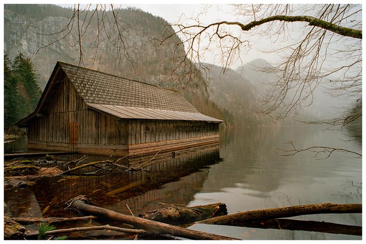 A wooden boathouse sits on a misty lake with mountains in the background and bare tree branches in the foreground.
