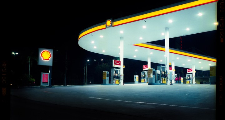 A brightly lit Shell gas station stands empty at night, with its distinctive yellow and red canopy illuminated.