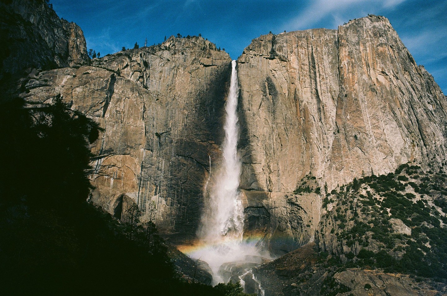 A tall waterfall cascades down a massive rock face, with a rainbow visible at its base.