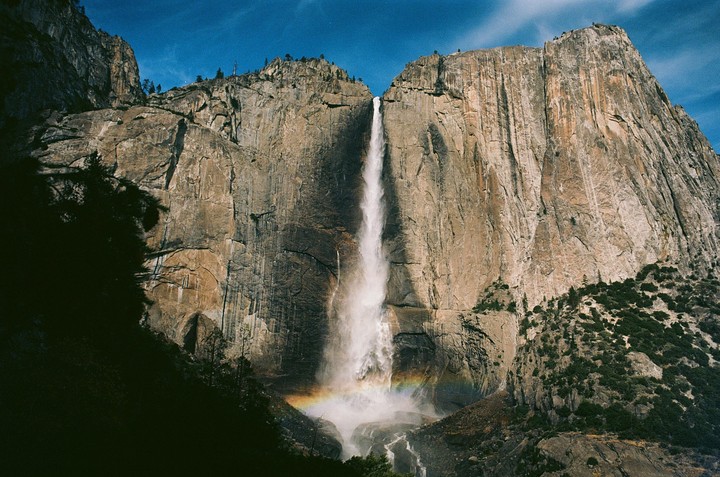 A tall waterfall cascades down a massive rock face, with a rainbow visible at its base.