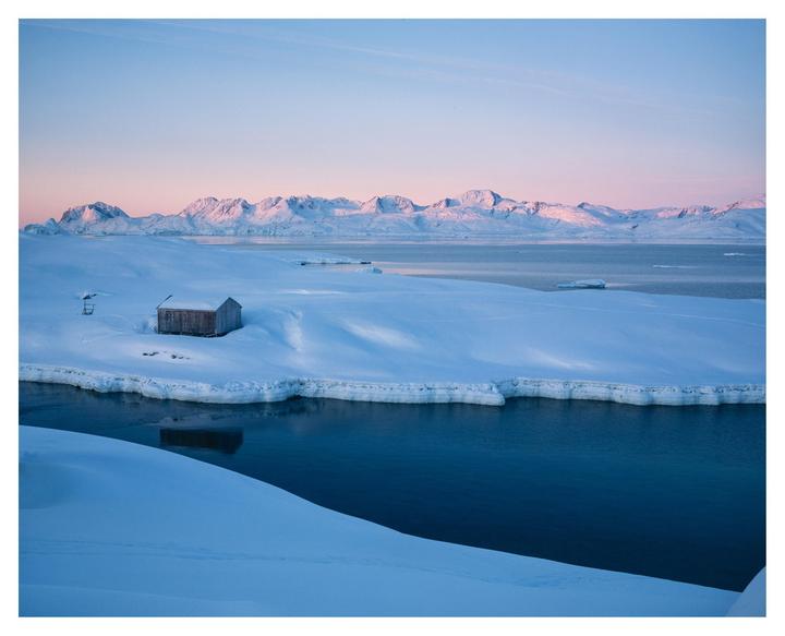 A small wooden cabin sits on a snow-covered bank next to a body of water, with mountains in the distance under a pink and blue sky.