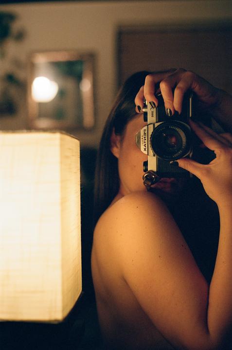 A woman takes a self-portrait in a mirror, holding a camera to her face, with a lamp illuminating her bare shoulder.