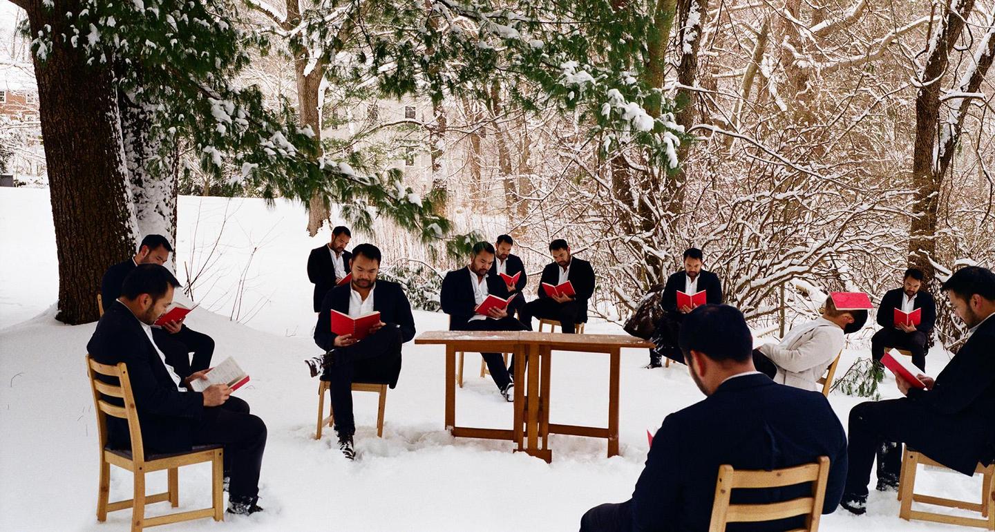 A group of men in suits are sitting on chairs in a snowy forest, reading books.