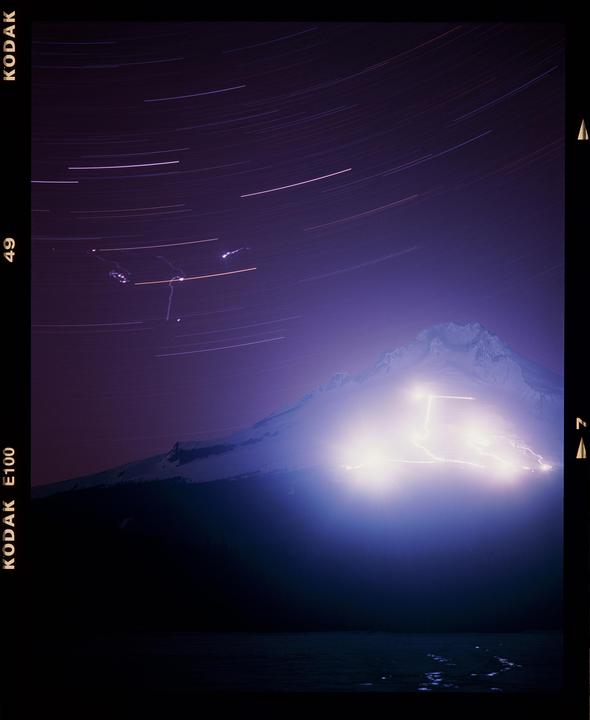 A mountain is illuminated by bright lights under a night sky with star trails.