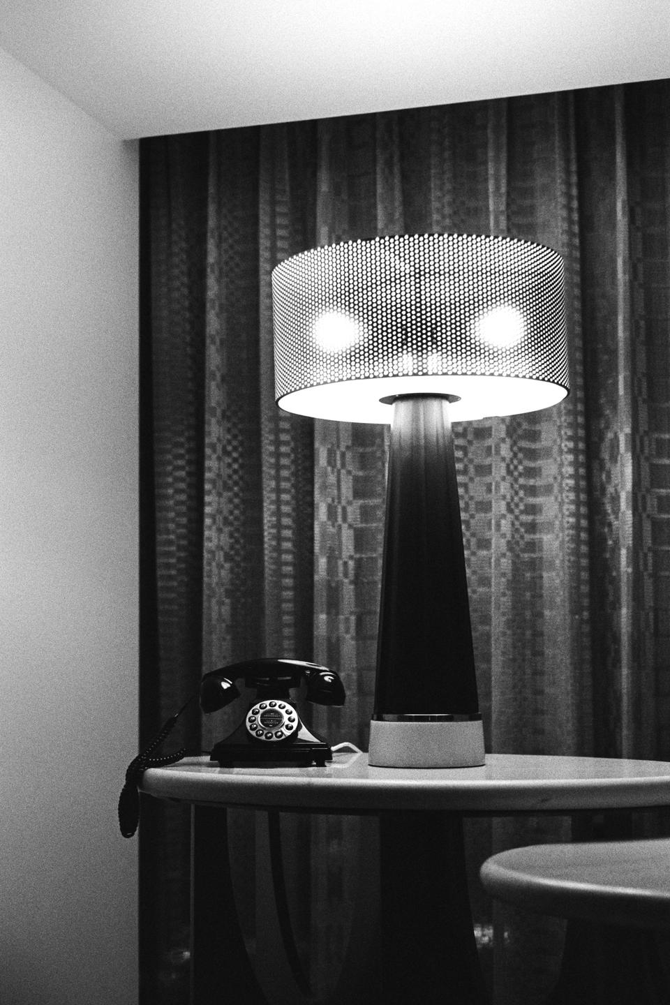 A rotary telephone and a lamp sit on a table in front of a patterned curtain.