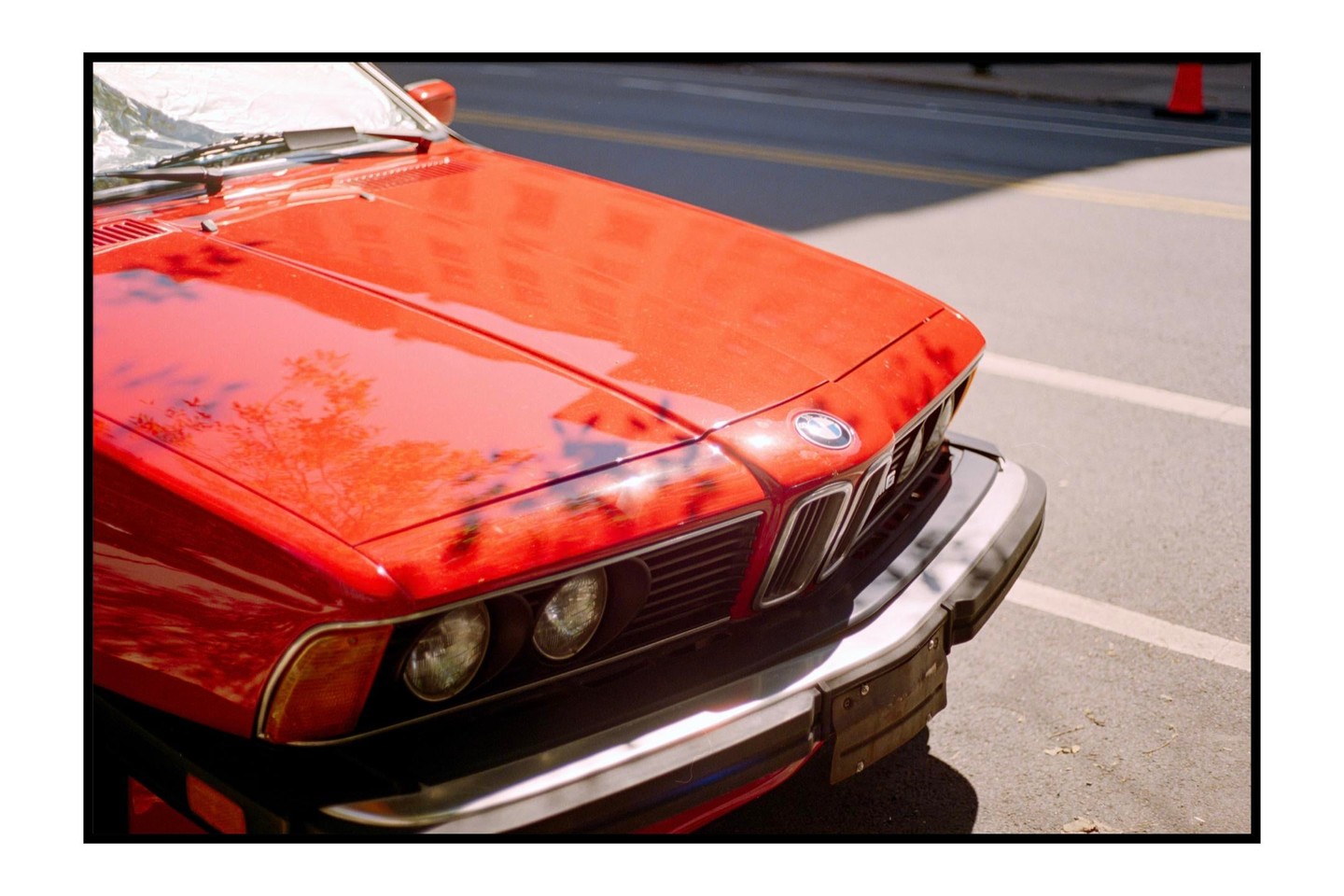 A red car is parked on an asphalt surface with tree reflections on its hood.