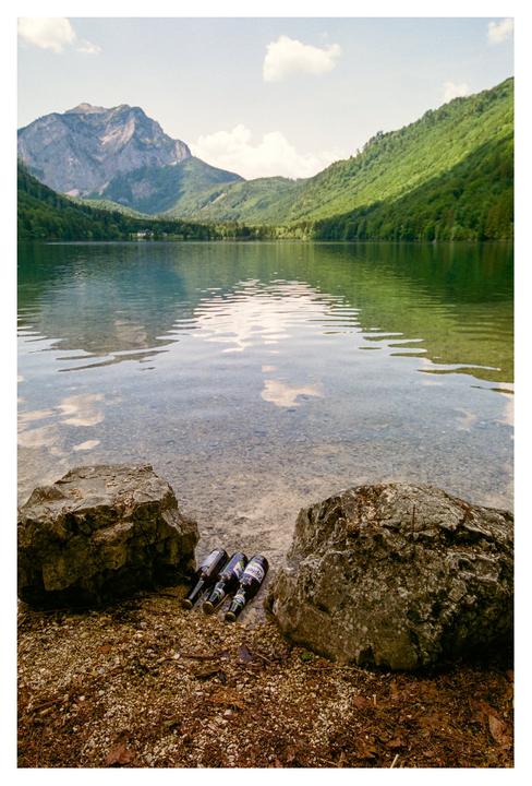 Three bottles rest on the rocky shore of a lake surrounded by mountains and forest.