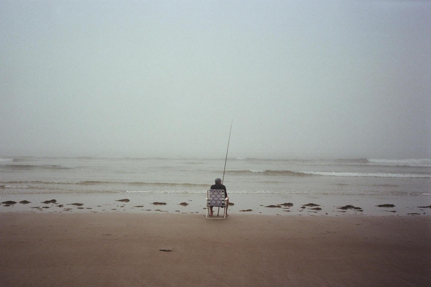 A person sits on a chair on a beach, fishing in the ocean on a hazy day.