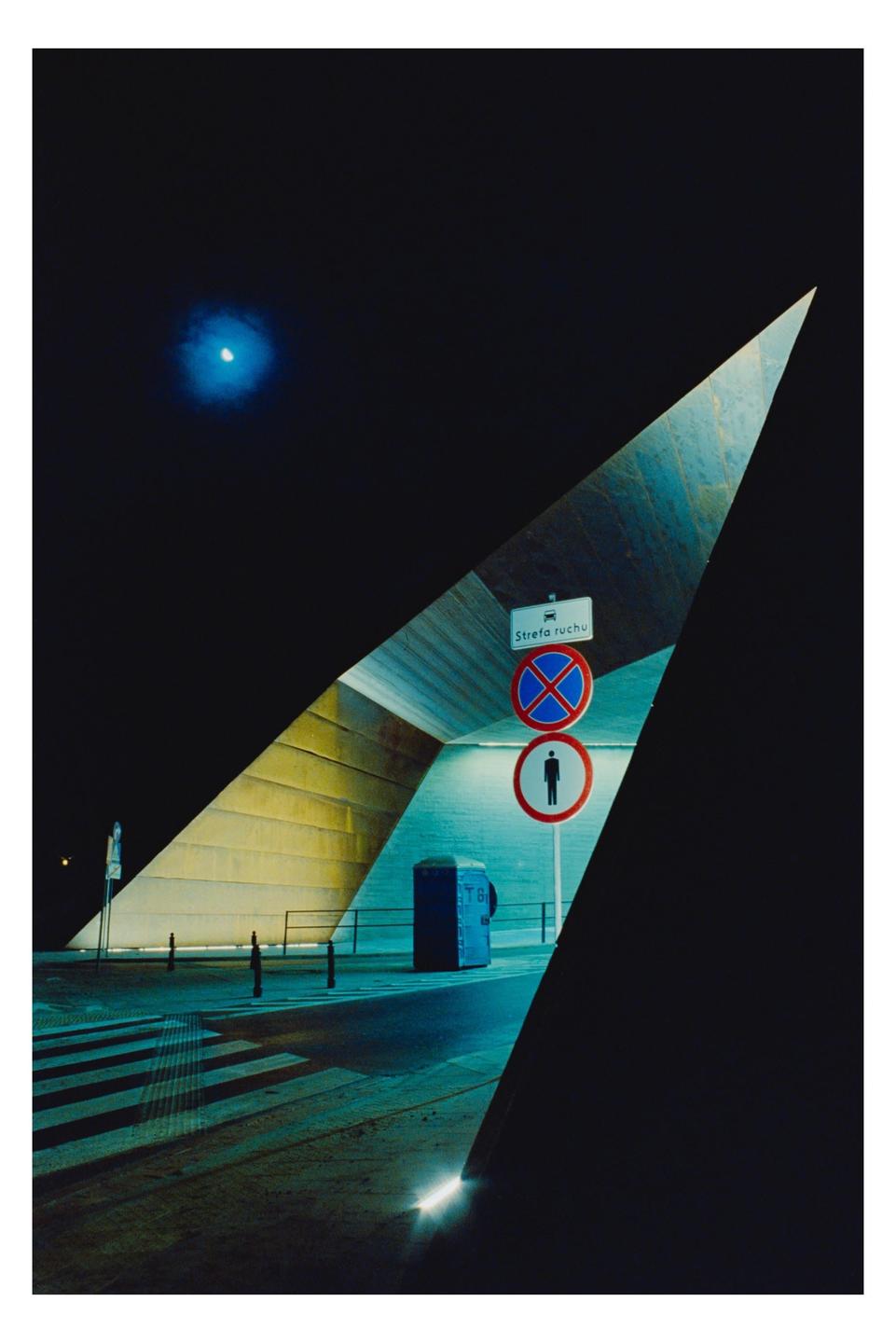 A blue moon shines over a street at night, with a crosswalk and street signs visible through a triangular opening.