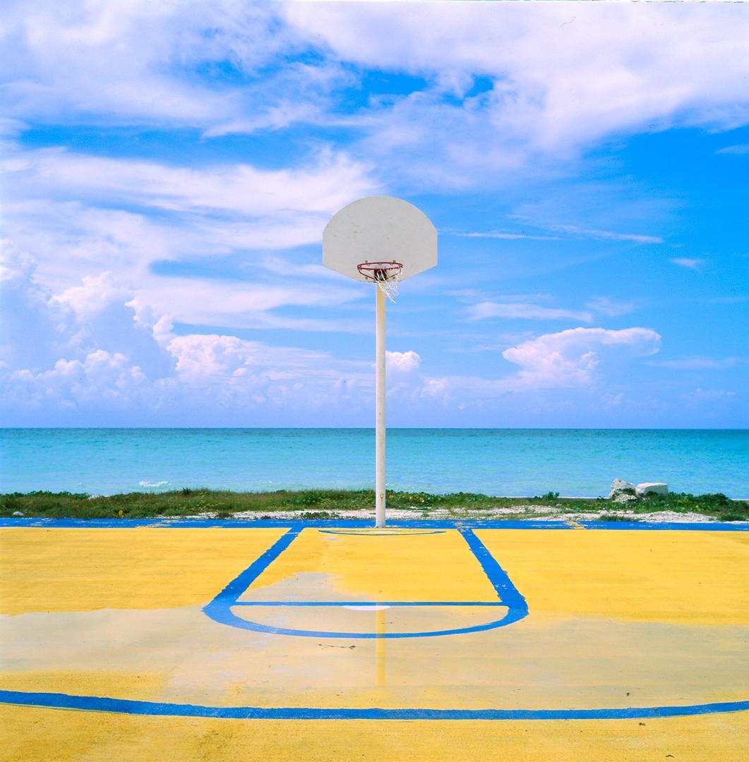 A basketball hoop stands on a yellow and blue court next to a turquoise ocean under a blue sky with clouds.