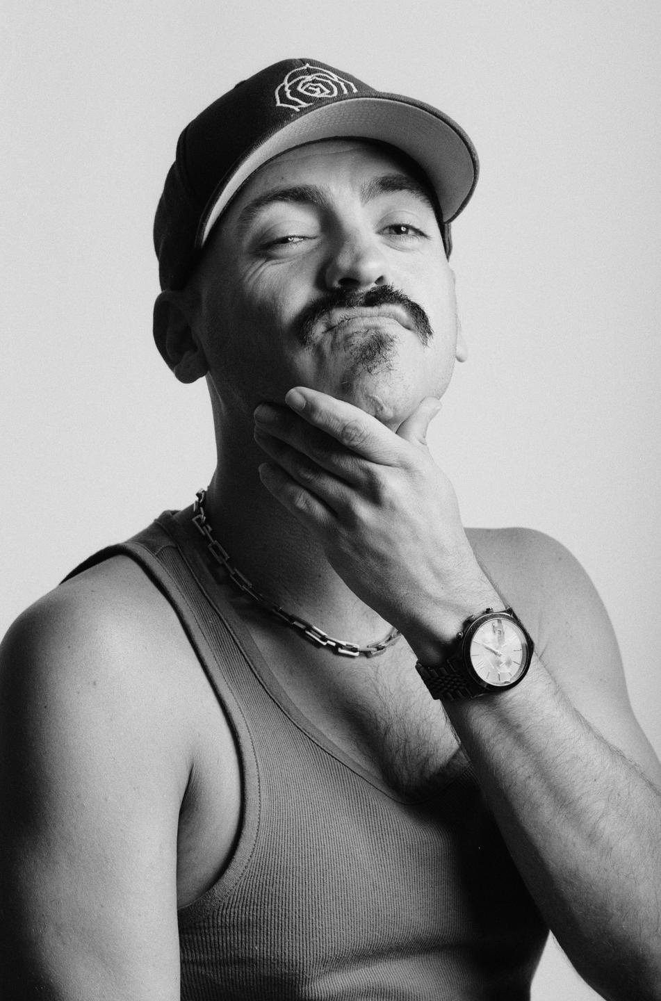A man with a mustache and a baseball cap poses with his hand on his chin.