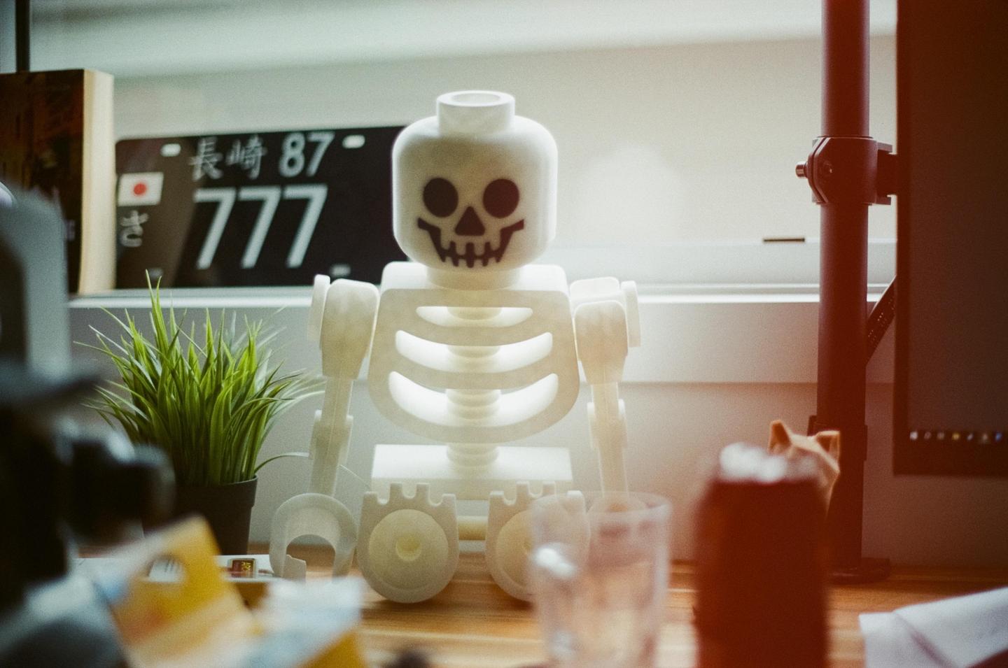 A white skeleton toy sits on a wooden surface next to a potted plant and a Japanese license plate.