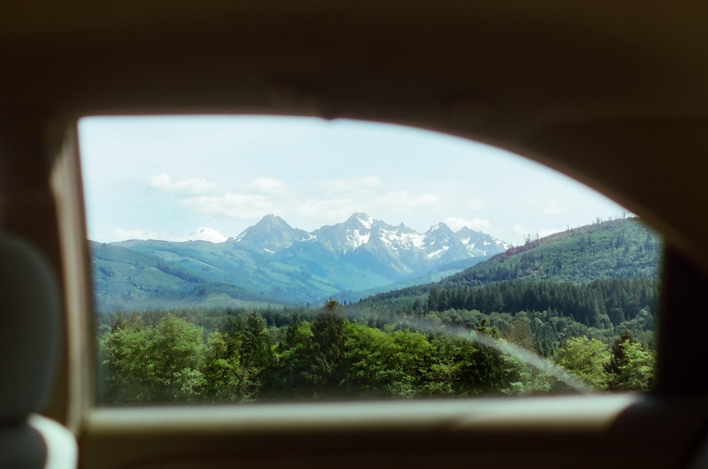 A view of snow-capped mountains and a forest is framed by a car window from the back seat.