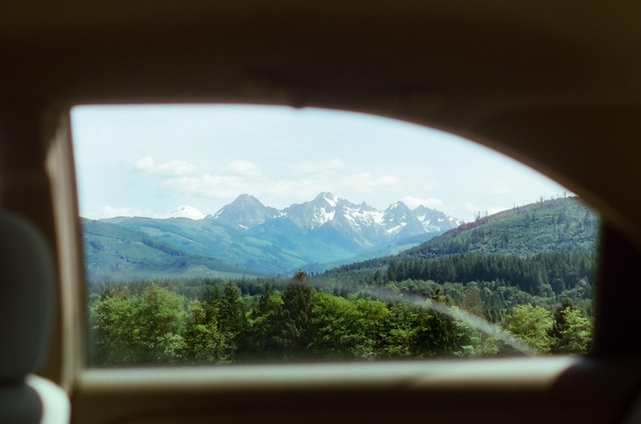 A view of snow-capped mountains and a forest is framed by a car window from the back seat.