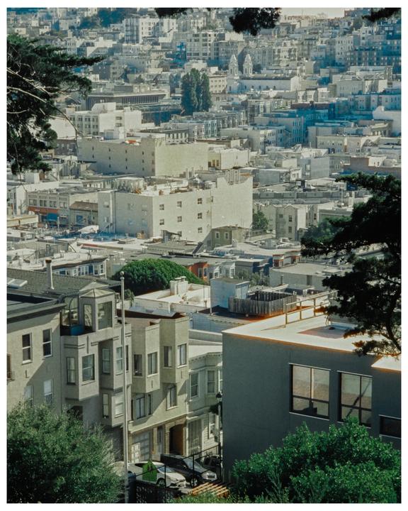 A hillside city is densely packed with buildings and trees under a bright sky.